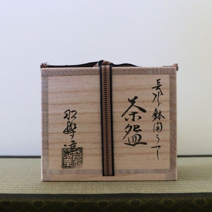 A light natural wood tea box with black Japanese calligraphy and a vertical black cord, displayed on a textured green mat against a plain light background.