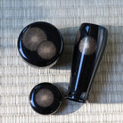 Three-piece glossy black lacquer set with metallic gold starburst patterns, including a tall vase, a round container, and a small lid, arranged on a woven straw mat.