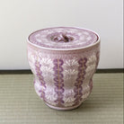 A decorative lidded ceramic jar with a purple and white floral pattern and gold accents, set on a light green woven mat against a plain light gray wall.