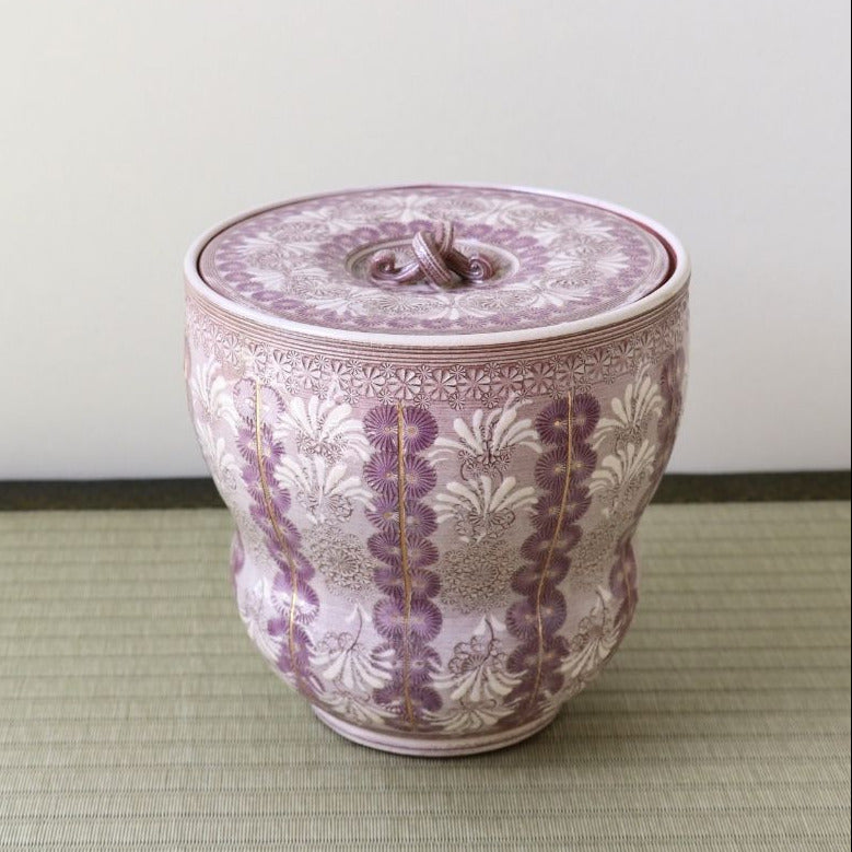 A decorative lidded ceramic jar with a purple and white floral pattern and gold accents, set on a light green woven mat against a plain light gray wall.