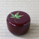 A round, glossy dark red lacquered box with a green maple leaf design and gold accents on the lid, set on a textured beige woven mat.