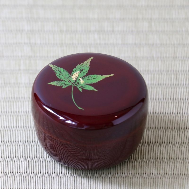 A round, glossy dark red lacquered box with a green maple leaf design and gold accents on the lid, set on a textured beige woven mat.