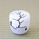 A small white cylindrical decorative box with a lid, decorated with a black bare tree, a yellow crescent moon, a haunted house, birds, and small cross motifs on a textured beige woven surface.