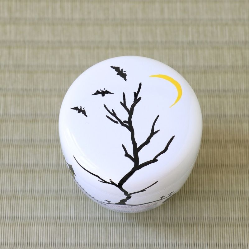 A small round white container or box lid decorated with a black bare tree, three black bats, and a yellow crescent moon, placed on a woven beige mat.