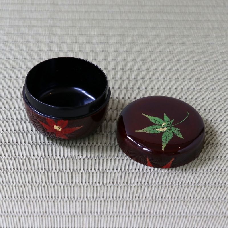 A glossy dark red lacquered box with its lid placed beside it on a woven light beige surface. The box body is black, and the lid and exterior are decorated with red maple leaf motifs and a green maple leaf with gold detailing.