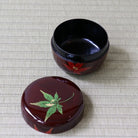 A glossy dark red lacquered round box with its lid placed beside it on a woven beige mat. The lid features a painted green maple leaf motif, and the open box shows a red floral design around the exterior.