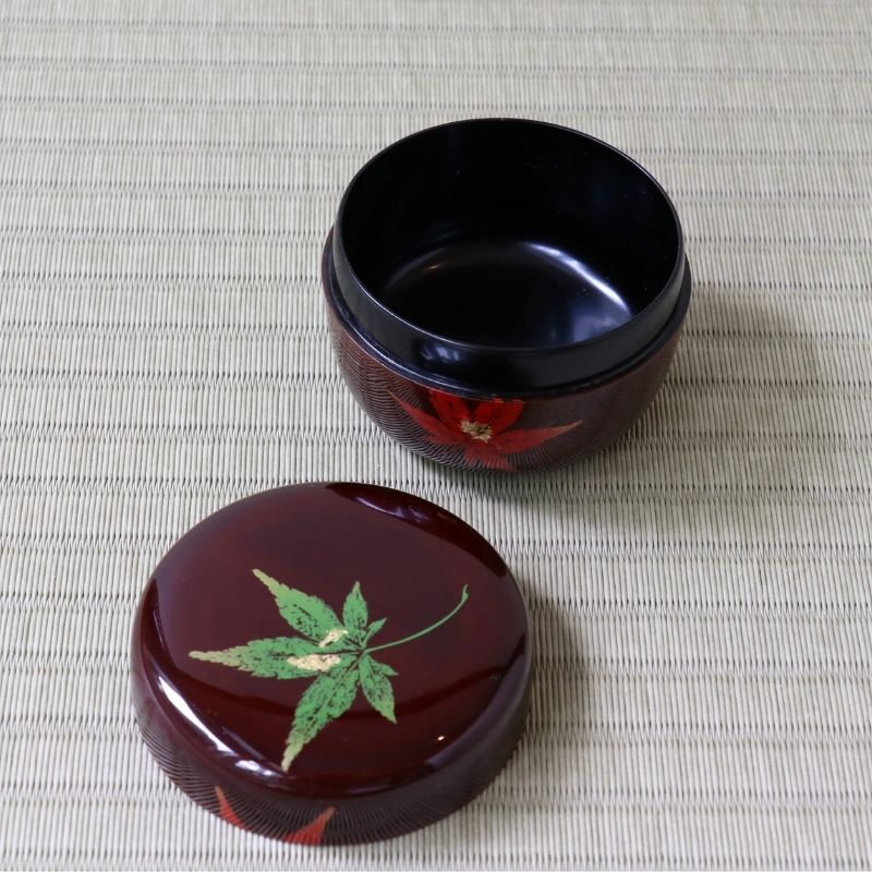 A glossy dark red lacquered round box with its lid placed beside it on a woven beige mat. The lid features a painted green maple leaf motif, and the open box shows a red floral design around the exterior.