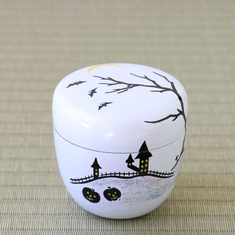 A small white round lidded container decorated with a Halloween night scene, featuring black bare tree branches, flying bats, a haunted house, fence, pumpkins, and a crescent moon, set on a beige woven mat background.