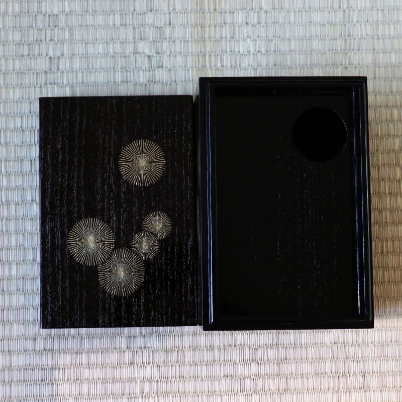 A pair of silver-toned sunburst earrings displayed on a black lacquered box next to its open black lid, set against a light beige woven straw mat background.