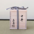 A light natural wood gift box with black Japanese calligraphy and a navy-and-white striped ribbon tied around it, placed on a woven mat against a plain light background.