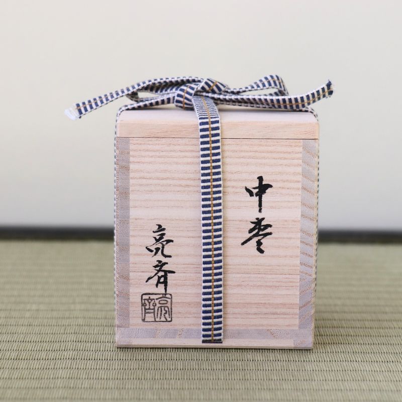 A light natural wood gift box with black Japanese calligraphy and a navy-and-white striped ribbon tied around it, placed on a woven mat against a plain light background.