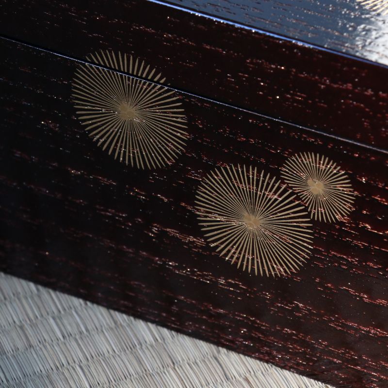 Close-up of a dark reddish-brown lacquered wooden box with three circular gold sunburst inlays, resting on a woven straw mat.