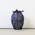 A small drawstring pouch made of dark navy blue patterned fabric with a purple cord tie, standing upright on a light textured surface against a plain off-white background.