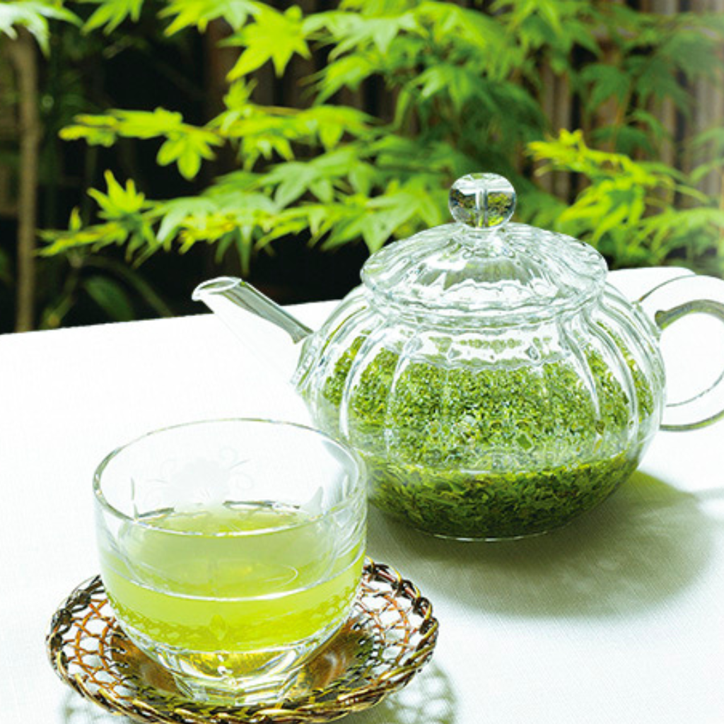 Clear glass teapot with green tea leaves and a glass of green tea on a white surface with greenery in the background.