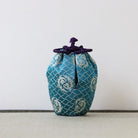 A small fabric drawstring pouch in teal blue with a white geometric pattern and a dark purple cord tie, standing on a light textured surface against a plain light gray background.