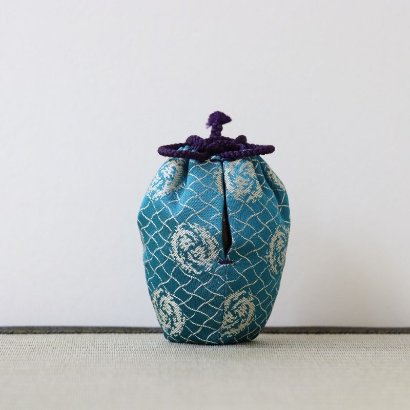 A small fabric drawstring pouch in teal blue with a white geometric pattern and a dark purple cord tie, standing on a light textured surface against a plain light gray background.