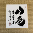 Black calligraphy on a white card with a gold border, placed on a woven mat.