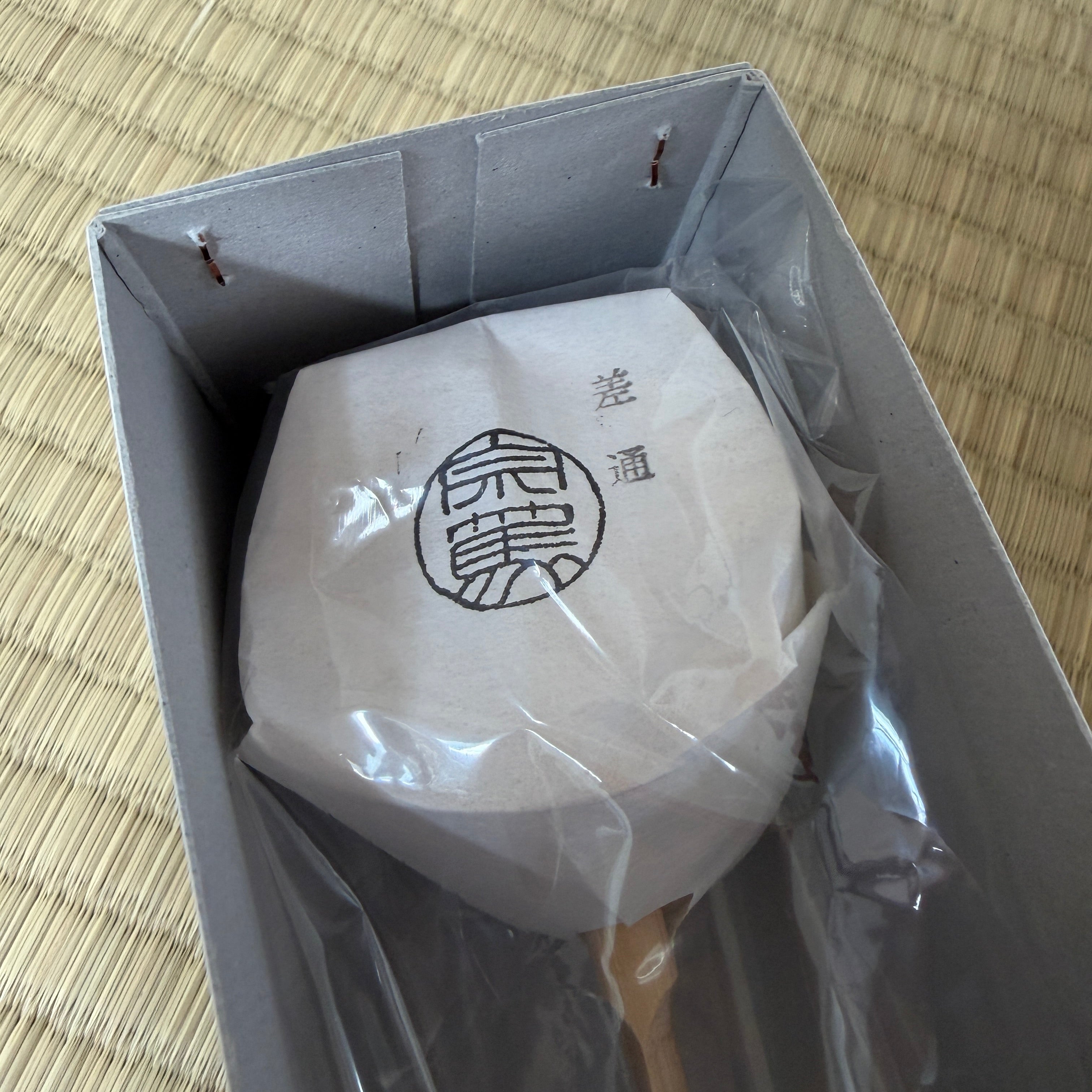 White product wrapped in plastic with a logo, inside an open box on a woven mat background