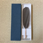 A dark brown feather quill with tiny white dots, displayed in a white presentation box next to its dark blue textured cover on a beige woven mat background.