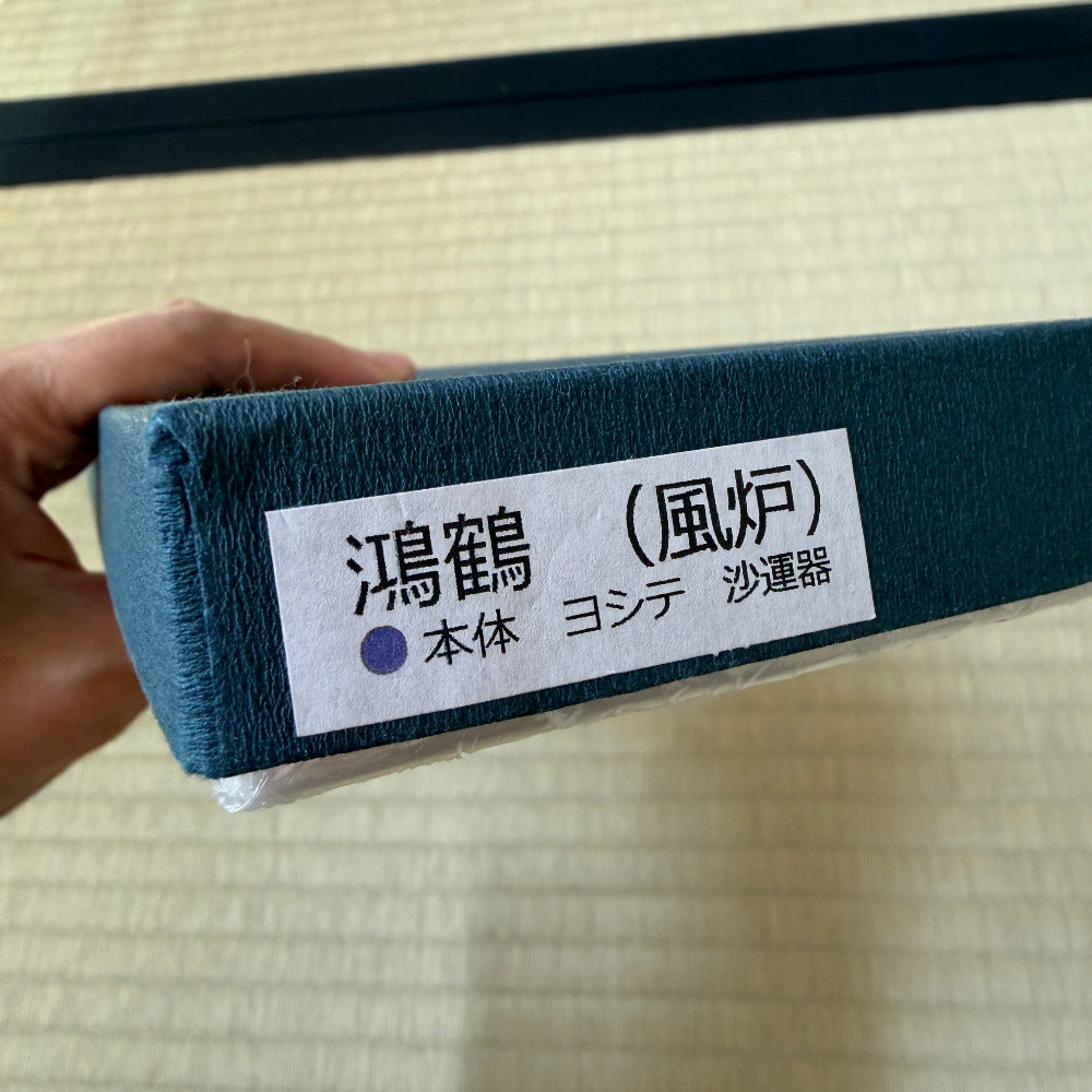 A hand holding a dark teal fabric-covered rectangular box with a white label printed in Japanese characters, set against a light beige textured background.