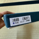 A hand holding a dark teal fabric-covered rectangular box with a white label printed in Japanese characters, set against a light beige textured background.