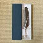 A traditional Japanese calligraphy ink feather quill with a brown feather and light wood handle, presented in a white box next to a dark navy blue textured cover on a beige woven mat background.