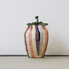 A small decorative fabric pouch or bag with vertical stripes in muted pink, cream, olive green, and purple, tied with a knotted dark green cord at the top, set against a plain light background.