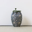 A small fabric drawstring pouch with a dark blue and beige floral brocade pattern and a green braided cord closure, standing on a textured surface against a plain light background.