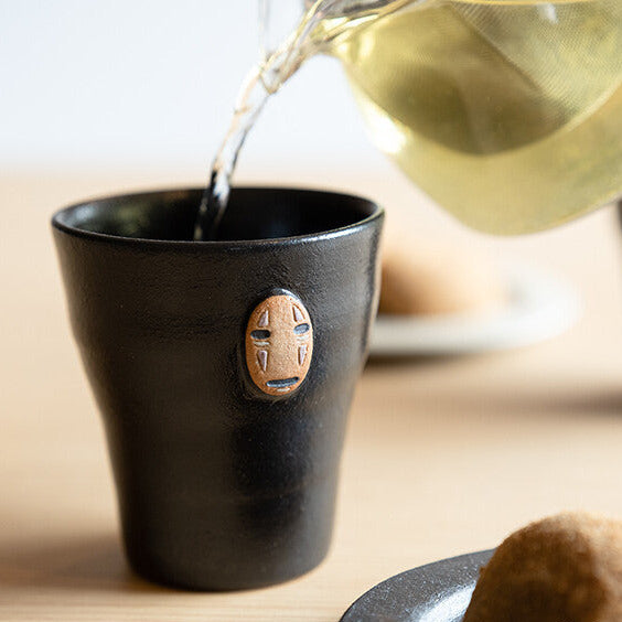 Black ceramic cup with a face design being filled with tea from a glass teapot.