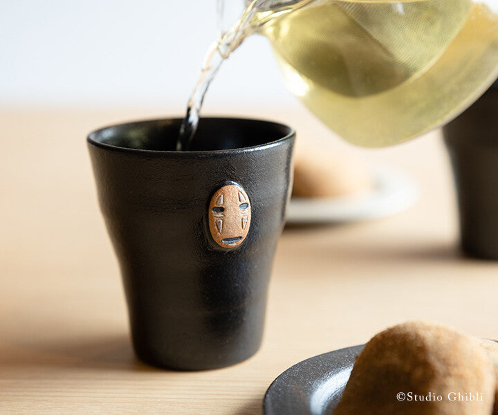 Black ceramic cup with a face design being filled with tea from a glass teapot.