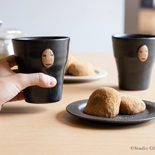 Two black ceramic cups with face designs on a wooden table with cookies.