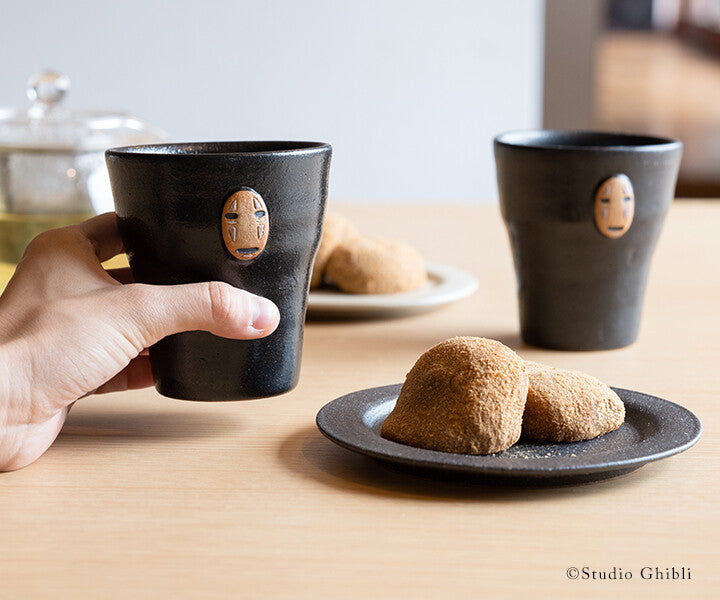 Two black ceramic cups with face designs on a wooden table with cookies.