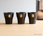 Three black ceramic cups with face designs on a wooden surface, featuring Studio Ghibli branding.