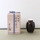 A light natural wood presentation box tied with a navy and white striped ribbon stands beside a dark amber-brown Takatori-glazed Katatsuki Chaire (Japanese shouldered tea caddy) with a white lid, set on a woven mat against a plain light wall.