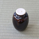 A small glossy dark brown ceramic jar with a white lid, placed on a light gray woven mat.