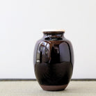 A glossy dark brown ceramic jar with a light beige lid, standing on a textured woven mat against a plain white background.