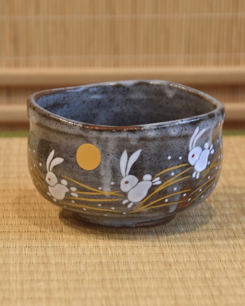 A traditional Japanese Kutani ware matcha bowl with a blue-gray glaze, featuring white rabbits and a golden moon motif on the exterior.