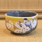 A Kutani Ware matcha bowl with a vibrant yellow exterior, featuring a detailed hand-painted design of a bird among cherry blossoms.