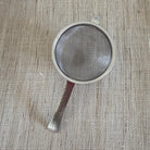 A small round metal mesh strainer with a hammered silver handle, placed on a textured beige burlap fabric background.