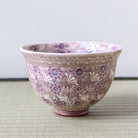 A vintage Japanese ceramic bowl decorated with a purple and beige floral pattern, resting on a woven mat against a neutral background.