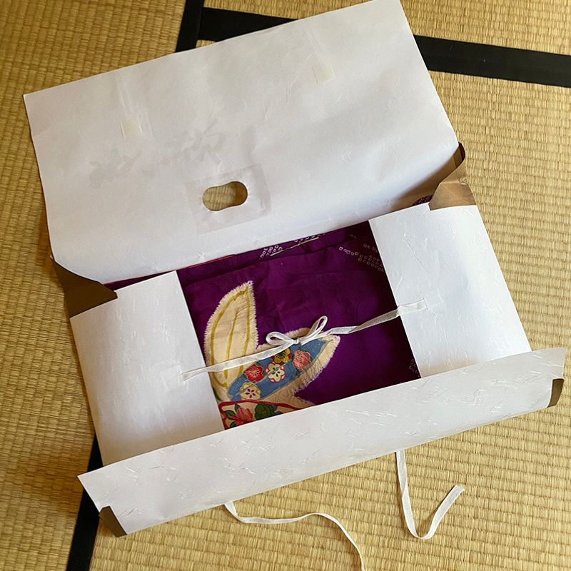 Nara Tea Co.'s set of 10 sheets of tatoushi paper Tatoushi l kimono Storage Paper with Strings, one of the product's packages is opened to reveal the knots in the interior strings and the pattern of a purple kimono.