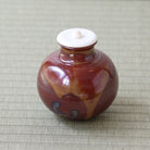 A small glazed ceramic lidded jar with a rounded body, featuring a marbled reddish-brown and tan glaze with dark accents, and a contrasting white lid, set on a woven beige mat.
