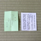 A folded light green booklet and a rectangular white card with Japanese text, placed on a woven straw mat background.