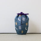A small fabric drawstring pouch in dark blue with white snowflakes and gold cherry blossoms, tied with a purple cord, standing on a light textured surface against a plain light gray background.