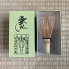 Bamboo whisk in a green box with Japanese text on a woven mat background