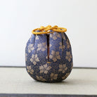 A small decorative fabric bag with a blue and beige floral pattern, featuring a drawstring closure at the top and a vertical slit, displayed on a textured surface against a plain light background.