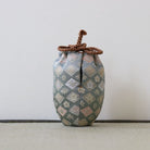 A small decorative fabric pouch with a diamond-patterned brocade design in muted blue, beige, and pink tones, featuring a brown braided drawstring closure at the top, set against a plain light gray background.
