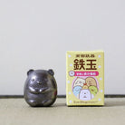 A small dark metallic Sumikkogurashi figurine stands next to its light yellow packaging box on a neutral surface against a light gray background.