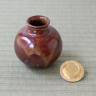 A small glazed ceramic chaire with a rounded body and short flared neck, shown next to its gold-toned lid on a woven beige mat. The vase has a glossy reddish-brown and amber marbled glaze.