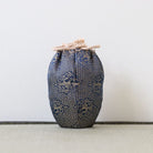 A small fabric drawstring pouch in dark blue and beige patterned material, standing upright against a plain light wall on a textured surface.